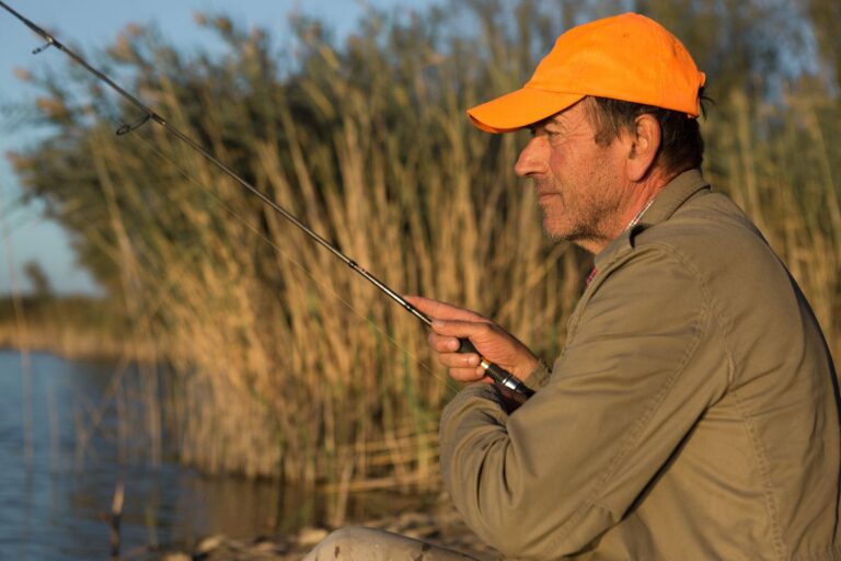 An old man fishing on a creek wearing orange cap.